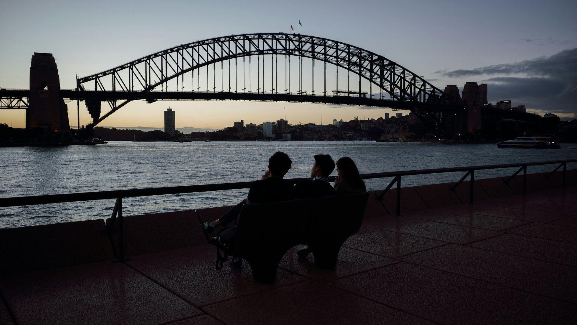 Three people on a bench watching Sydney Harbour Bridge at dusk